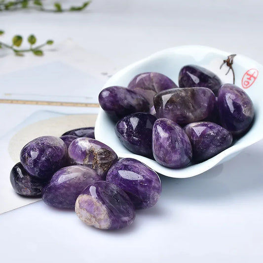 Amethyst Tumble Stone Purple crystals on a white surface with a white dish containing more crystals.