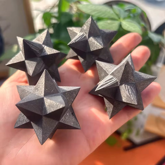 Shungite Merkaba Star Four dark metallic star-shaped throwing stars held in a person's hand, with a blurred indoor background featuring green plants.
