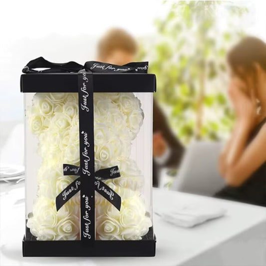 White Rose Teddy Bear White rose heart-shaped bouquet in a clear box with a black ribbon, set against a blurred background of two people sitting at a table.