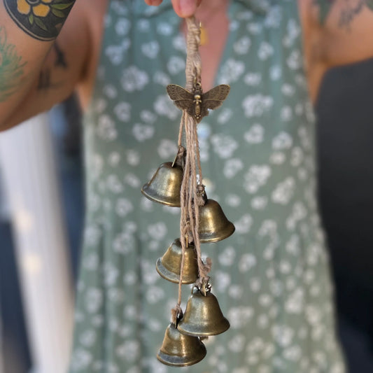 Witches Protection Bells A close-up of a person holding a hanging string of five small brass bells with a butterfly ornament at the top. The person is wearing a sage green floral dress, and the background is softly blurred.