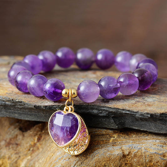 Amethyst Healer Bracelet with Heart Charm Purple beaded bracelet with a gold heart-shaped pendant on a stone surface