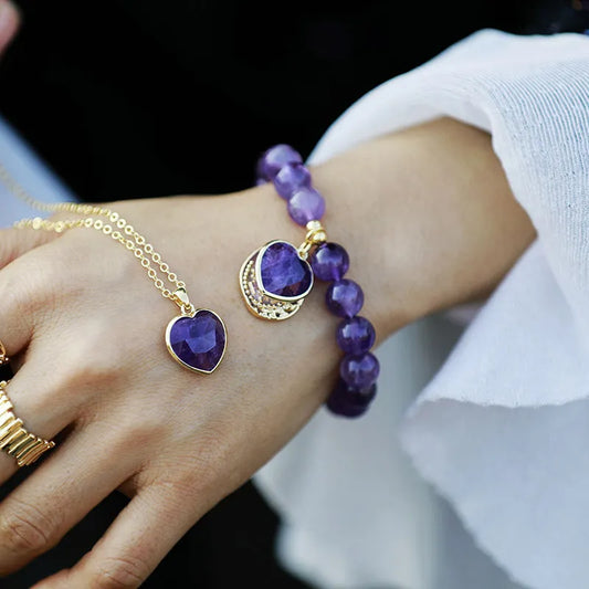Amethyst Healer Bracelet with Heart Charm A close-up of a person's hand and wrist wearing jewelry: a gold chain necklace with a faceted purple heart pendant, a gold ring, and a purple beaded bracelet with a matching heart charm.