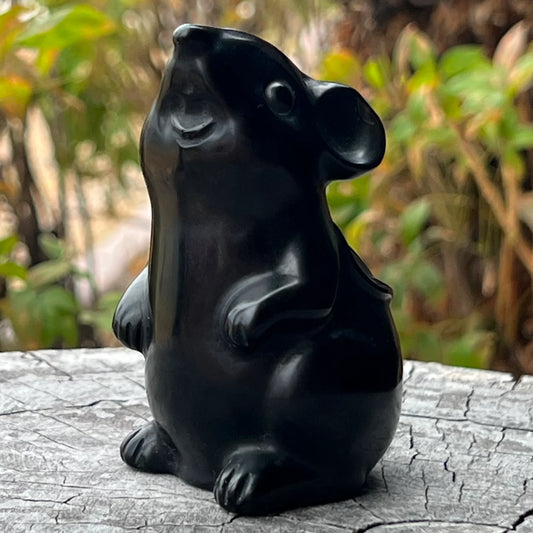 Black Obsidian Mouse A glossy black mouse figurine sitting upright on a weathered gray log with a blurred green and brown natural background.