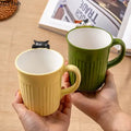 Cute 3D Cat Hanging Mug green and beige variant front side GiftDealAU product image. Two mugs, one yellow and one green, held by a person on a wooden surface with a magazine in the background.