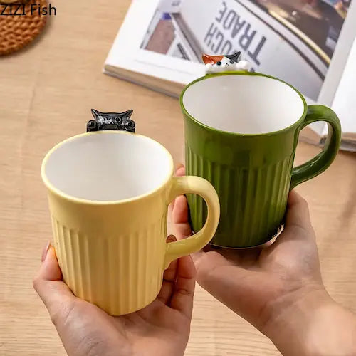 Cute 3D Cat Hanging Mug green and beige variant front side GiftDealAU product image. Two mugs, one yellow and one green, held by a person on a wooden surface with a magazine in the background.