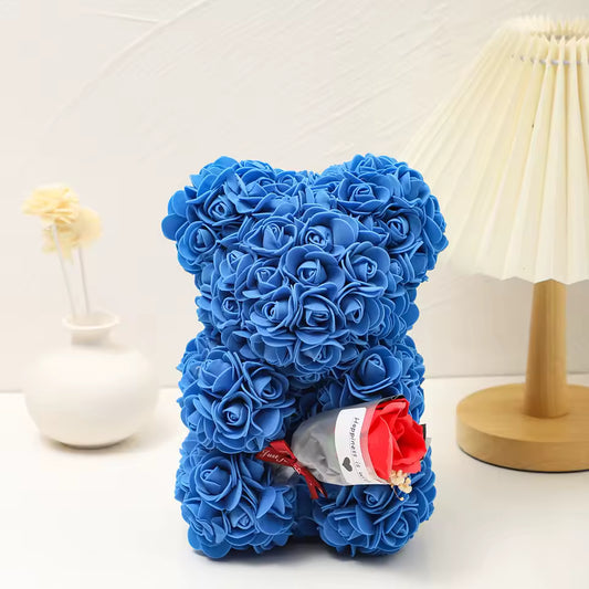 Foam Rose Teddy Bear Holding Bouquet Blue teddy bear made of roses on a white surface with a lamp and vase in the background.