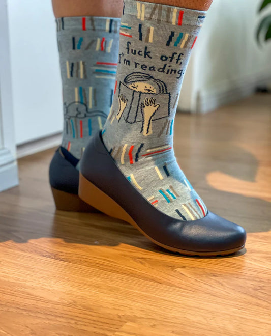 Gray crew socks with a book-themed pattern and the text 'fuck off, i'm reading' worn with dark wedge shoes on a wooden floor.