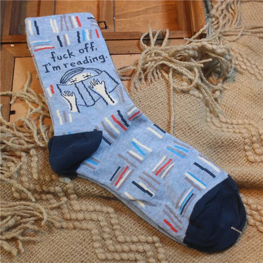 Fuck Off I'm Reading Socks A single light blue crew sock with a navy heel and toe, featuring a printed graphic of hands holding an open book with the text 'fuck off, I'm reading:' and vertical book spines in red, blue, white, and navy, displayed on a woven jute rug.