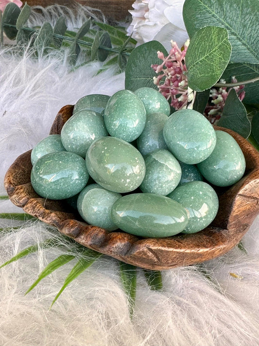 Green Aventurine Tumble Stones A pile of smooth, polished light green stone eggs arranged in a small carved wooden bowl, styled with green leaves and white fluffy feathers.