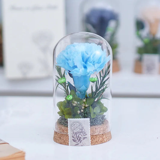 Preserved Flower in Glass Dome Blue flower in a glass dome with plants on a white surface