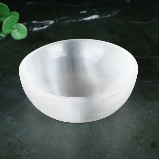 Selenite Cleansing & Charging Bowl A frosted glass bowl with a smooth, matte finish, shown on a dark textured surface with a small green leaf in the top-left corner.