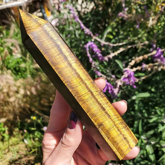 Tiger Eye Crystal Tower A hand holding a polished golden-yellow crystal tower with natural striated patterns, set against a blurred outdoor background of green foliage and purple flowers.
