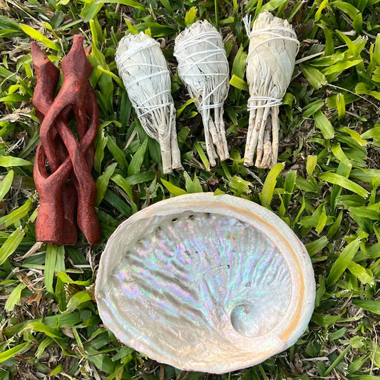 A set of natural crystals and herbs arranged on green grass, including three white sage bundles tied with string, a reddish-brown carnelian tree piece, and a large iridescent abalone shell.