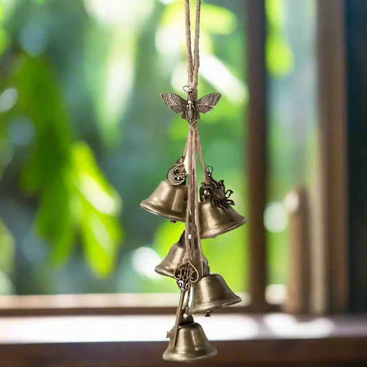 Witches Protection Bells A hanging decorative wind chime with three small metallic bells suspended from a string, featuring a butterfly ornament at the top, photographed in front of a softly blurred window background.