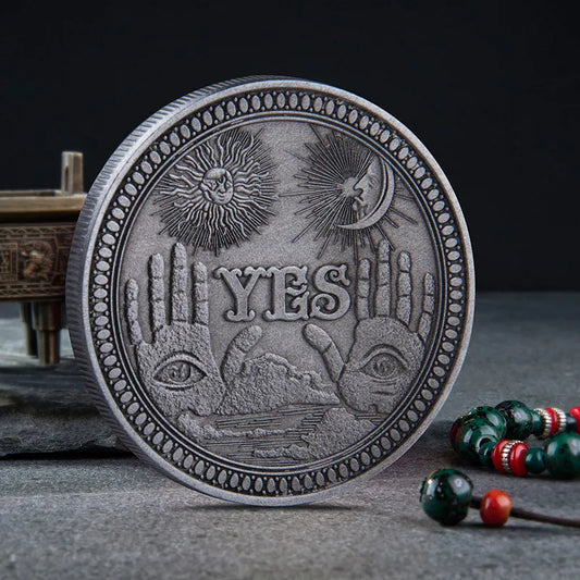 Yes No Boxed Divination Decision Coin A round silver-toned metal coin or medallion with embossed religious and symbolic imagery, including the word 'YES' across the center, two hands with eyes, a sun, a crescent moon, and a decorative border. It is photographed on a dark textured surface with a blurred decorative object and beaded strand in the background.
